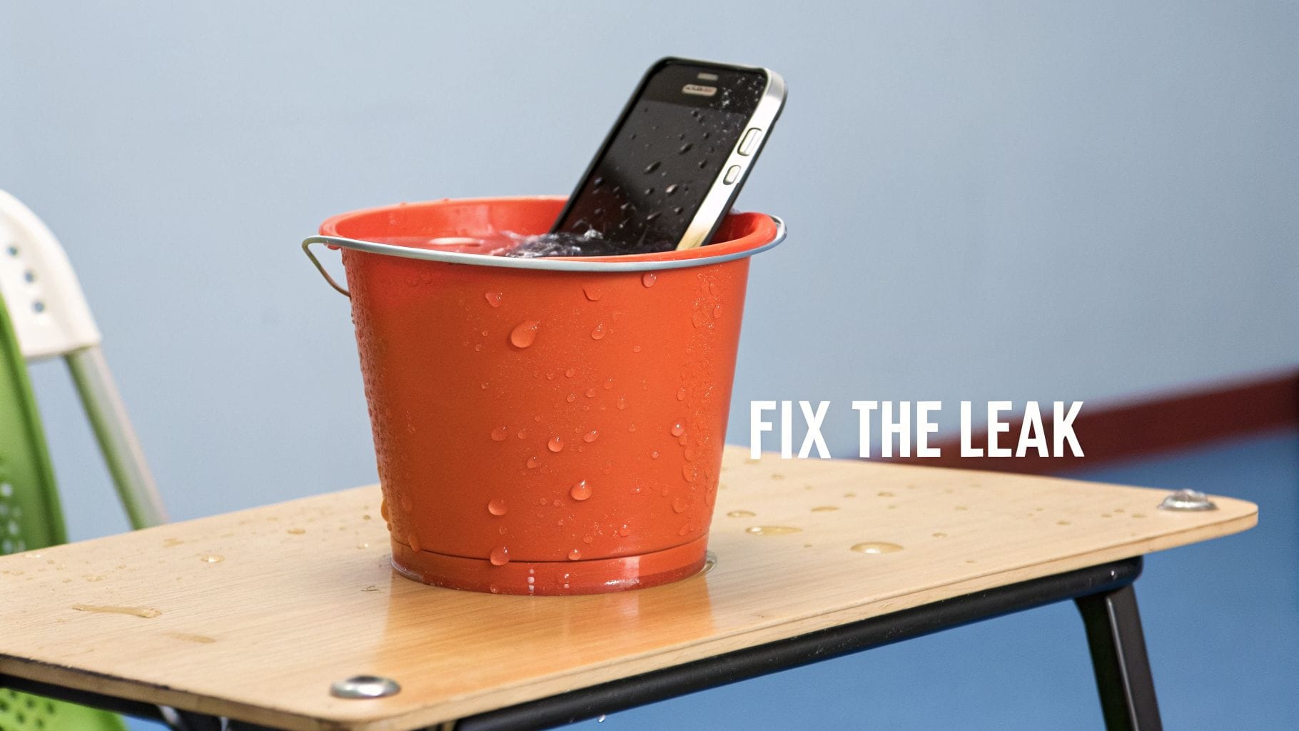 A smartphone partially submerged in an orange bucket of water on a wooden desk, with 'FIX THE LEAK' text.